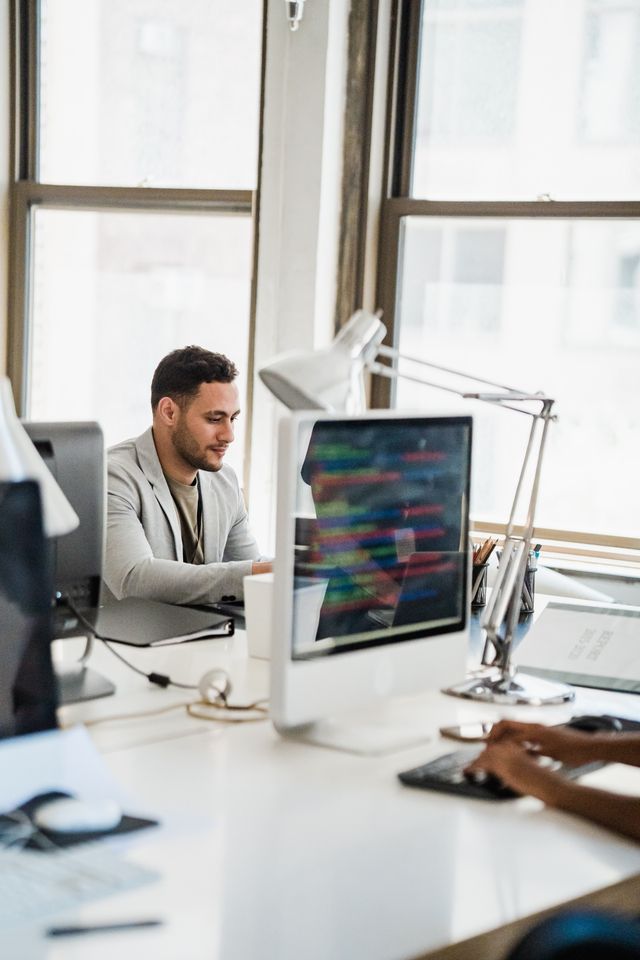 Man in blazer working on a business presentation at desktop computer setup near window in modern office studio fot. PEXELS